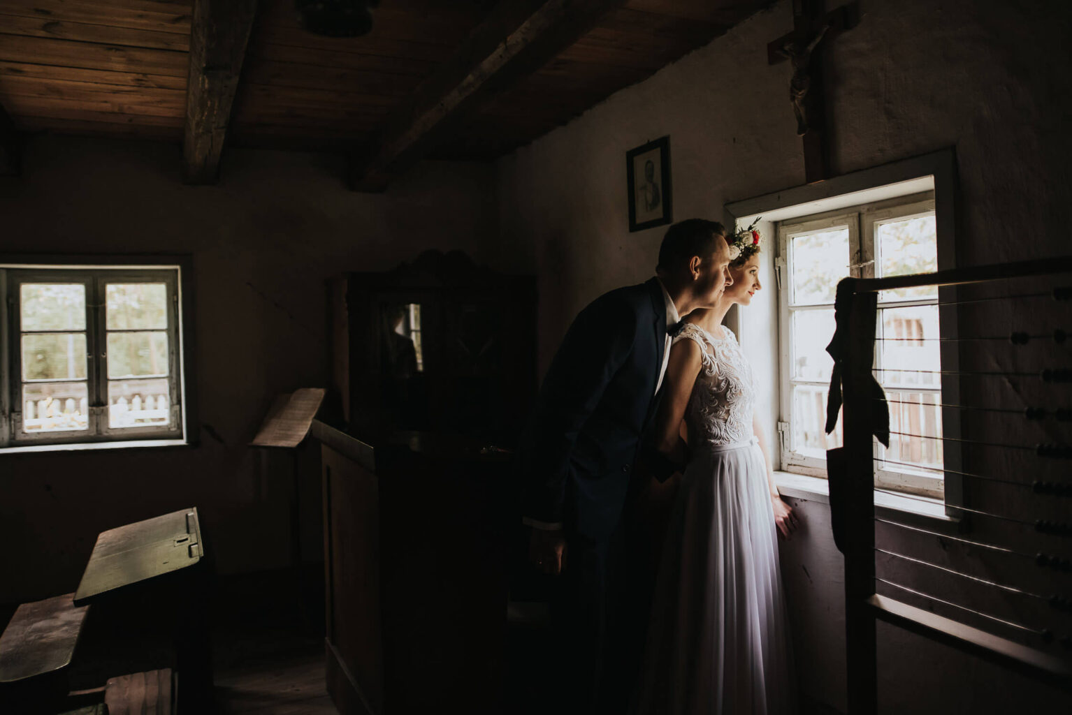 Para młoda wygląda przez okno w skansenie podczas rustykalnego pleneru ślubnego