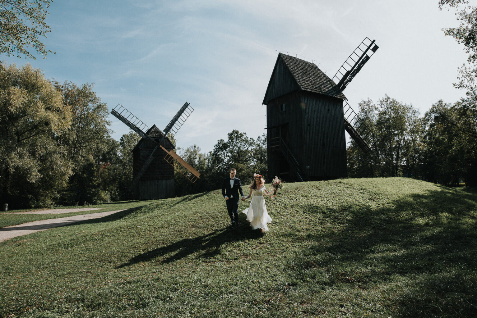 Kadr z rustykalnej sesji zdjęciowej przedstawiający zbiegająca ze wzgórza parę młodą, na tle wiatraków Muzeum wsi opolskiej.